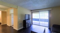 This single-floor space offers a clean, functional layout designed for comfortable everyday living. The main living area is a standout feature, showcasing rich dark hardwood flooring that contrasts beautifully with the warm yellow-toned walls and a textured ceiling overhead. A full-width sliding glass door floods the room with abundant natural light and opens to an outdoor balcony, offering pleasant views of surrounding greenery. Vertical blinds provide flexible light control and privacy. The living area provides ample space for seating and entertainment arrangements, with a built-in media setup area that accommodates a flat-screen television. A floor vent indicates a central HVAC system for year-round climate comfort. An adjacent area, visible through a doorway, hints at additional rooms that contribute to the overall flow of the space. The overall ambiance is bright, airy, and welcoming, with a practical design that balances open living with functional details. The combination of natural light, outdoor access, and thoughtful layout makes this a versatile and inviting space suited for a variety of lifestyles.