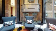 This striking space masterfully merges historic and contemporary design elements across its single floor. Exposed brick walls, remnants of the original structure, form a dramatic backdrop throughout, juxtaposed against sleek white polished floors and warm wood paneling. The entry area features floor-to-ceiling glazing, flooding the space with natural light while offering views of the surrounding streetscape. Rich walnut sliding panels and dark metal accents frame the transition between zones, creating a refined sense of arrival.

A lounge area showcases high-backed upholstered chairs in deep grey, positioned atop a vibrant abstract area rug, complemented by sculptural side tables. Intricate metalwork screens and curated planters add organic texture against the aged brick backdrop. A dedicated workstation hub sits centrally on a circular patterned rug, furnished with modern computer equipment and red upholstered seating, illuminated by cascading pendant lighting above.

A communal dining area features a live-edge wood table paired with bold yellow upholstered chairs and anchored by a dramatic black marble base. The soaring atrium ceiling with glass panels draws in abundant natural light, unifying the space with an airy, gallery-like ambiance throughout.