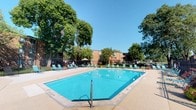 This outdoor amenity area centers around a large, rectangular swimming pool with clear turquoise water, offering a refreshing and inviting atmosphere during warm-weather months. The pool deck is finished in a clean concrete surround and is furnished with multiple lounge chairs positioned along the perimeter, providing ample space for sunbathing and relaxation. A shaded gazebo structure at one end of the deck offers a covered retreat from direct sunlight, adding functional variety to the outdoor space. The area is enclosed by a perimeter fence, ensuring privacy and security for residents. Mature deciduous trees frame the space generously, providing natural shade and a lush, park-like ambiance that softens the surrounding brick residential buildings. The open sky above allows for abundant natural light throughout the day. Overall, the pool area is well-maintained and thoughtfully arranged, creating a communal outdoor environment that balances recreational use with comfort. The surrounding greenery and classic brick architecture contribute to a grounded, established character, making this outdoor space a notable feature of the community.
