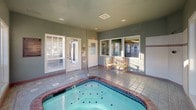 This single-level indoor spa room is purpose-built for relaxation and leisure. The centerpiece of the space is a generously sized hot tub with a classic brick-tiled surround, fitted with stainless steel handrails for safe entry and exit. The jet-equipped basin holds clear, blue-green water and is set into a spacious tiled floor that extends throughout the room.

The room is enclosed by soft sage-green painted walls accented with white tile wainscoting, creating a clean and calming atmosphere. Large, multi-pane white-framed windows line multiple walls, allowing abundant natural light to pour in while offering views of the surrounding exterior grounds and neighboring structures. This bright, sunlit quality gives the room an open and airy feel despite being fully enclosed.

Seating is provided by a solid wooden bench positioned along the wall, offering a comfortable spot to rest before or after soaking. The room is equipped with clearly marked exit doors and informational signage posted near the entryways, ensuring guest safety and convenience. Overhead, recessed lighting and a central HVAC vent contribute to year-round climate comfort. Overall, the space provides a well-maintained, functional, and inviting environment ideal for unwinding and relaxation.