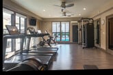Located at 10210 N Foothill Blvd in Cupertino, CA, this fitness center is a thoughtfully designed, single-room facility that maximizes its compact footprint with a comprehensive selection of exercise equipment. The space features light hardwood-style flooring throughout, neutral-toned walls, and recessed lighting complemented by dual ceiling fans that ensure consistent air circulation during workouts.

The cardio zone is well-stocked with multiple treadmills, elliptical machines, and stationary cycling bikes — including Peloton-style spin bikes — each equipped with individual display screens. A dedicated strength training area houses a multi-function cable machine and a functional trainer board, while a full rack of dumbbells lines one wall alongside weight benches, catering to a wide range of resistance training needs.

Large framed windows along one wall flood the space with natural light and offer direct views of an outdoor swimming pool, creating an energizing atmosphere. Wall-mounted televisions are positioned throughout for entertainment during workouts. Full-length mirrors span a significant portion of one wall, visually expanding the space and providing form-monitoring capability. A wall-mounted air conditioning unit ensures climate control year-round. The overall design is clean, functional, and well-organized, making it an efficient and inviting fitness facility.