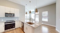 This single-floor space showcases a contemporary design with a cohesive neutral color palette of soft grays and warm whites throughout. The open-concept layout connects the kitchen and living area seamlessly, creating a spacious, airy feel enhanced by abundant natural light from multiple windows dressed with clean white blinds.

The kitchen is a standout feature, equipped with white shaker-style cabinetry extending to the ceiling, granite countertops, and a full suite of stainless steel appliances including a French door refrigerator, range, microwave, and dishwasher. A peninsula island with an undermount sink serves as both a functional workspace and casual dining area, illuminated by a trio of brushed gold pendant lights. A light blue subway tile backsplash adds a subtle design accent.

The living area features warm hardwood-style flooring, a ceiling fan, and a sliding door providing outdoor access. Bedrooms offer plush carpeting and generous natural light. Both bathrooms are finished with matching granite vanities, white cabinetry, and hardwood-style flooring, with the primary bath including a walk-in closet and combination tub-shower. The overall ambiance is clean, modern, and move-in ready.