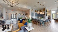 This thoughtfully designed single-floor clubhouse presents an open-concept layout that seamlessly connects multiple functional zones, creating a cohesive and inviting communal environment. The space is anchored by soaring double-height ceilings and an abundance of natural light, courtesy of oversized windows and clerestory glazing that frame direct views of the outdoor pool and amenity area.

The kitchen is a standout feature, outfitted with full-height slate-blue shaker cabinetry accented by brushed gold hardware, granite countertops, a large island with bar seating, stainless steel appliances, and a decorative herringbone tile backsplash. Globe pendant lights and ornamental gold wall medallions lend a bold, curated aesthetic throughout.

Adjacent lounge areas are furnished with a mix of warm leather seating, upholstered sofas in mustard and cobalt tones, and woven accent chairs arranged around coffee tables with gold-framed bases. A stone-clad accent wall with a mounted television and built-in display shelving anchors one seating zone. A separate high-top dining area with round tables and metal-framed stools sits beside the pool-facing windows. Throughout, large-format tile flooring and carefully selected décor create a polished, resort-inspired atmosphere.