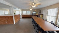 This single-floor facility is thoughtfully designed to accommodate gatherings and community activities across several distinct functional areas. The entry lobby sets a welcoming tone with terracotta tile flooring, recessed lighting, and a textured ceiling. Large glass doors flood the space with natural light and provide views of the outdoor surroundings, while a seating area with a floral-patterned sectional sofa and an accent chair offers a comfortable place to relax or wait.

Moving deeper into the space, a generously sized lounge area continues the carpeted flooring and features an expansive built-in sectional sofa lining the walls, accented by framed artwork and windows dressed with vertical blinds that allow abundant natural light. A central fireplace with a decorative surround and built-in shelving serves as a focal point, adding warmth and character to the room.

The meeting and dining area is anchored by a long rectangular conference-style table surrounded by numerous chairs, making it well-suited for group meetings or community events. Dual ceiling fans ensure comfortable air circulation throughout. Along one wall, warm wood cabinetry with a countertop provides a functional kitchenette or serving area, complete with small appliances and a sink. Large windows along this wing further enhance the bright, open atmosphere of the space.