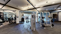 Located at 2000 Taylor St in Houston, TX, this fitness center offers a generously sized, single-floor exercise space designed to support a full range of workout routines. The room features durable rubber tile flooring throughout, providing both safety and comfort underfoot. Architectural interest is added by the distinctive X-shaped exposed beam detail on the ceiling, complemented by recessed lighting and ceiling fans that keep the space well-lit and ventilated.

The cardio zone lines one wall with a row of treadmills and elliptical machines, each equipped with individual screens, all positioned near large windows that invite natural light and outdoor views. A dedicated free weights area includes a full dumbbell rack and an adjustable bench, while a cable cross machine and power rack with weight plates support comprehensive strength training. Wall-mounted flat-screen televisions are positioned throughout for entertainment during workouts.

The walls are adorned with bold geometric mural artwork in a gray-and-white palette, adding a dynamic visual energy to the space. Full-length mirrors along one wall enhance the sense of openness and allow for form checks during exercise. Glass entry doors provide a clean, modern entryway. The overall ambiance is polished and motivating, balancing functional fitness infrastructure with thoughtful, contemporary design.