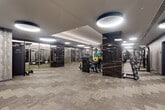 Located at 1425 New York Ave NW in Washington, DC, this premium fitness center spans a generous single floor, offering a well-appointed and thoughtfully designed workout environment. The space is divided into distinct zones, including an open main gym floor, a glass-enclosed studio, and a dedicated cardio and strength training area.

The interior design strikes a sophisticated tone, with richly veined dark marble-clad columns, textured wood-effect wall panels, and a patterned carpet floor that adds visual depth throughout. Overhead, large circular pendant lights are recessed into a tiered ceiling, providing bright, even illumination across the entire facility.

The main gym floor is generously proportioned, housing a wide array of strength machines, free weights, and benches. A dedicated free-weight zone features a full rack of color-coded bumper plates alongside multiple cable and resistance machines. The cardio section is equipped with treadmills, stair climbers, and other cardio machines, each outfitted with personal screens.

The glass-enclosed studio, framed in gold-toned hardware, offers a separate training environment with turf flooring, ideal for functional fitness or group sessions. Floor-to-ceiling mirrors line multiple walls throughout the space, amplifying the sense of openness and allowing for form monitoring during workouts. The overall ambiance is modern, refined, and purpose-built for high-performance training.