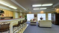 This single-floor space offers a warm and functional layout designed to encourage relaxation and social engagement. The main area is anchored by a comfortable lounge zone furnished with neutral-toned sofas accented with decorative pillows, arranged to promote conversation and ease. Adjacent to the lounge, a raised counter with elegant wrought iron bar stools provides an additional seating option, ideal for casual gatherings or informal meetings.

The interior features warm olive-toned walls complemented by crisp white wainscoting and decorative crown molding, lending the space a polished, classic character. Recessed lighting and overhead fluorescent fixtures ensure the space is well-lit throughout. Multiple windows fitted with white blinds allow natural light to filter in, brightening the carpeted lounge area while contributing to an open and airy atmosphere.

Decorative touches such as potted plants and framed artwork on the walls add personality and a welcoming feel to the environment. The flooring transitions between tile near the counter area and plush carpet in the lounge, subtly defining each functional zone. Overall, this space strikes a balance between practical amenities and comfortable design, making it well-suited for everyday use and community interaction.