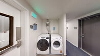 This shared laundry area, located at 16210 NE 80th St in Redmond, WA, is a compact, efficiently organized utility space designed to serve building residents. The room is neatly finished with white walls and a combination of hard flooring near the appliances and blue-grey carpeting in the adjacent corridor area.

At the heart of the space sit two Whirlpool front-load appliances — a washer and a dryer — positioned side by side beneath a visible ventilation duct that runs vertically to the ceiling. The machines are clearly labeled and accompanied by posted instructional signage for resident use.

The room also houses essential building utility infrastructure, including an electrical panel and an adjacent warning-labeled utility box mounted on the wall, ensuring centralized access to key systems. A smoke detector is visible on the ceiling, reflecting attention to safety standards.

Access to the laundry area is provided through a solid-core door featuring a narrow vertical window and a stainless steel push bar, connecting directly to Stair N1. An illuminated green exit sign is mounted above the doorway, reinforcing the space's compliance with safety codes. The overall design is clean, functional, and straightforward, prioritizing practicality for everyday residential use.