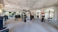 Located at 500 Trinity Lane North in Saint Petersburg, Florida, this fitness center combines upscale architectural design with a comprehensive suite of exercise equipment. The single-floor space is distinguished by soaring coffered and groin-vaulted ceilings adorned with crown molding, lending a refined, almost residential elegance rarely found in a fitness facility. Tall arched windows and French doors line the walls, flooding the interior with abundant natural light and offering views of the lush surrounding landscape and outdoor pool area. The flooring features durable fitness-grade tile throughout, providing a clean and functional workout surface. The space is well-equipped for a full-body training experience, offering a multi-station cable and Smith machine, a full dumbbell rack, adjustable benches, resistance machines, and an array of stability balls and resistance accessories. Wall-to-wall mirrors along one side visually expand the room and assist with form and technique. Mounted flat-screen televisions provide entertainment during workouts. A built-in cabinetry unit with a beverage refrigerator adds a thoughtful amenity. The overall ambiance is bright, open, and polished, making this fitness center a standout facility that seamlessly blends architectural sophistication with practical, high-quality functionality.