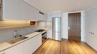 Located at 1201 Half St SE in Washington DC's Capitol Riverfront neighborhood, this modern single-floor residence showcases a clean, contemporary design with high-quality finishes throughout. The open-concept living area is bright and airy, featuring light hardwood flooring and large floor-to-ceiling windows that invite abundant natural light and offer views of the surrounding urban environment. The kitchen is thoughtfully appointed with flat-front white cabinetry, warm wood accent shelving with under-cabinet lighting, sleek quartz countertops, and stainless steel appliances including a gas range with oven and a full-size refrigerator. The entry is framed by a distinctive warm wood door, adding a striking design contrast to the otherwise crisp, white interior palette. The bathroom continues the modern aesthetic with a floating walnut vanity topped with a grey concrete-style countertop, an undermount sink, a frameless glass walk-in shower, and a large backlit mirror, all set against clean white walls and neutral tile flooring. Throughout the residence, the consistent use of light tones, natural wood accents, and minimalist detailing creates a cohesive and sophisticated living environment.