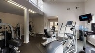 Located at 1100 Tunnel Road in Asheville, North Carolina, this single-floor fitness center offers a thoughtfully designed workout space combining functional layout with an inviting atmosphere. The facility is divided into distinct workout zones, each tailored to different types of exercise.

The main cardio area features soaring vaulted ceilings with a wood-paneled ceiling detail and pendant lighting, creating an airy, open feel. Multiple windows along the walls allow abundant natural light to fill the space, while a wall-mounted television provides entertainment during workouts. A water cooler station adds everyday convenience. French doors at one end of the room open directly to an outdoor deck, enhancing ventilation and connecting the interior to the natural surroundings.

A dedicated strength training area houses multiple commercial-grade cable and resistance machines, including Batca-brand equipment, arranged to allow comfortable movement between stations. Structural columns define this zone while maintaining visual openness throughout.

Durable rubber flooring covers the entire facility, providing cushioning and traction suited for a high-use fitness environment. The neutral gray walls and clean white trim throughout contribute to a bright, professional aesthetic. The overall design balances practicality with comfort, making this a well-rounded fitness facility.