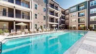 This multi-story residential community is designed around a vibrant, amenity-rich courtyard that serves as the centerpiece of the property. Spanning two floors and beyond, the building's exterior showcases a modern architectural palette, blending neutral stucco tones with dark board-and-batten siding and accents of teal and green, creating a visually dynamic façade. Private balconies extend from multiple levels, offering residents elevated views of the lushly landscaped courtyard below.

The outdoor amenity area is anchored by a generously sized swimming pool with crystal-clear blue water, bordered by a wide tiled sun deck lined with cushioned lounge chairs and shade umbrellas — ideal for relaxation and recreation. Adjacent to the pool, a covered outdoor terrace provides a comfortable gathering space, furnished with high-top tables and chairs beneath a pergola-style structure with recessed lighting, perfect for casual socializing or al fresco dining.

Tropical palm trees and carefully curated greenery are integrated throughout the courtyard, lending a resort-like ambiance to the space. A secondary pool area features umbrella-shaded seating and additional patio furnishings, further enhancing the outdoor living experience. The community's cohesive design and thoughtfully appointed amenities create an inviting, upscale atmosphere throughout.