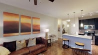 This well-appointed single-floor residence offers an open-concept layout that seamlessly connects the living area to the kitchen, creating a spacious and inviting atmosphere. The living room is anchored by rich hardwood flooring and features a ceiling fan with track lighting overhead, lending a contemporary feel to the space. A plush leather sofa, complemented by decorative throw pillows, sits beneath a striking pair of large abstract canvas artworks in warm amber and brown tones, adding a touch of artistic character to the room. Neutral, light-toned walls throughout enhance the sense of openness and allow natural light to circulate freely. The kitchen area is defined by a raised breakfast bar with stone countertops and pendant lighting hanging above, creating an ideal casual dining or entertaining space. Bar stools tuck neatly under the counter, while the sink and cabinetry are finished in clean white, maintaining a bright, modern aesthetic. A mirrored accent and decorative wall art in the transitional hallway add depth and visual interest. The cohesive design blends warm textures with modern fixtures, resulting in a comfortable yet refined living environment suitable for everyday living and entertaining alike.
