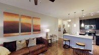 This well-appointed single-floor residence offers an open-concept layout that seamlessly connects the living area to the kitchen, creating a spacious and inviting atmosphere. The living room is anchored by rich hardwood flooring and features a ceiling fan with track lighting overhead, lending a contemporary feel to the space. A plush leather sofa, complemented by decorative throw pillows, sits beneath a striking pair of large abstract canvas artworks in warm amber and brown tones, adding a touch of artistic character to the room. Neutral, light-toned walls throughout enhance the sense of openness and allow natural light to circulate freely. The kitchen area is defined by a raised breakfast bar with stone countertops and pendant lighting hanging above, creating an ideal casual dining or entertaining space. Bar stools tuck neatly under the counter, while the sink and cabinetry are finished in clean white, maintaining a bright, modern aesthetic. A mirrored accent and decorative wall art in the transitional hallway add depth and visual interest. The cohesive design blends warm textures with modern fixtures, resulting in a comfortable yet refined living environment suitable for everyday living and entertaining alike.