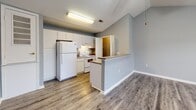 Located at Caraway Commons in Jonesboro, Arkansas, this one-bedroom, one-bathroom unit offers a practical and well-designed single-floor layout. The open-concept kitchen and living area forms the heart of the home, featuring durable wood-look vinyl flooring throughout. The kitchen is equipped with white cabinetry, a white electric range, a full-size refrigerator, and a double stainless steel sink with laminate countertops. A peninsula-style breakfast bar separates the kitchen from the living area, creating a natural flow between the two spaces. A ceiling fan is installed in the living area, and natural light filters in through a large window. The bedroom is a comfortable, well-proportioned room finished with soft carpet flooring and multiple windows fitted with blinds, allowing for natural light and outdoor views. The bedroom connects to a walk-in closet with wire shelving for organized storage and carpet underfoot. The bathroom features wood-look vinyl flooring, a tub/shower combination, a toilet, a single vanity with cabinetry, and accessibility-friendly grab bars. Neutral gray walls carry throughout the unit, complemented by white trim and textured ceilings, giving the space a clean, cohesive appearance.
