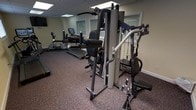 This single-floor fitness room offers a functional and well-organized workout space designed to accommodate a variety of exercise needs. The room is outfitted with a prominent multi-station strength training machine at its center, featuring cable pulley systems, adjustable seats, and multiple attachment points for a full range of resistance exercises. A treadmill with an integrated display screen is positioned along the back wall, complemented by additional cardio equipment nearby, providing options for both aerobic and strength-based training.

The space features durable, speckled carpet flooring well-suited to the demands of a fitness environment. Neutral-toned walls contribute to a clean, distraction-free atmosphere, while white-trimmed windows with slatted shutters allow natural light to filter into the room without compromising privacy. A drop ceiling with recessed lighting panels ensures consistent, even illumination throughout the space.

The layout is efficiently arranged to maximize usability without overcrowding, allowing comfortable movement between equipment. The overall ambiance is practical and purpose-driven, making this fitness room a convenient and capable space for regular exercise routines.