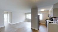This single-floor space offers a clean, modern aesthetic defined by its open-concept layout and neutral color palette. The living area is spacious and airy, featuring light hardwood-style flooring and crisp white baseboards that contrast gently against the warm beige walls. Natural light flows in through multiple windows, keeping the interior feeling fresh and welcoming throughout the day.

A structural column subtly divides the living space from the kitchen area, maintaining an open feel while providing a natural sense of separation. The kitchen is equipped with a stainless steel refrigerator and benefits from its own window, allowing for additional natural light. Flush-mount ceiling fixtures provide consistent overhead lighting across both areas.

The overall ambiance is calm and understated, with a minimalist design that offers a versatile canvas for a variety of interior styles. The continuity of flooring materials across both spaces reinforces the cohesive, unified character of the layout. Clean lines, ample wall space, and thoughtful use of natural light make this a functional and inviting environment.