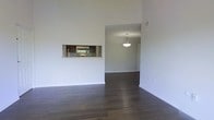 This single-floor space makes a strong first impression with its dramatic, soaring ceilings that amplify the sense of openness throughout the main living area. Rich, dark hardwood flooring runs continuously across the space, grounding the clean, neutral aesthetic established by the crisp white walls and trim.

A defining architectural feature is the built-in pass-through window that connects the kitchen to the main living area, offering both visual connectivity and functional convenience. The kitchen, glimpsed through the pass-through, is outfitted with dark cabinetry and stainless steel appliances, including an over-the-range microwave, set against a light countertop that provides an appealing contrast.

An open doorway leads into an adjoining room, where a pendant light fixture serves as a subtle focal point, hanging elegantly from the ceiling and casting a warm glow. The continuity of the hardwood flooring throughout ties the spaces together, creating a cohesive and unified flow from room to room.

The overall ambiance is clean, modern, and understated, with an emphasis on open space and natural proportions. The neutral color palette and minimal detailing make the space highly adaptable, offering a versatile canvas suited to a variety of interior styles and furnishing preferences.