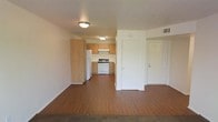 This single-floor unit offers an open-concept layout that seamlessly connects the living area to the kitchen, creating a sense of spaciousness despite its compact footprint. The flooring throughout is a warm, medium-toned hardwood-style surface that adds visual continuity across the main living space.

The kitchen is positioned at the far end of the open area and is outfitted with light natural wood cabinetry, providing ample storage. White appliances, including a refrigerator and a freestanding range with an overhead ventilation hood, are neatly integrated into the layout. The kitchen receives overhead lighting via a flush-mounted ceiling fixture, keeping the space well-lit.

The living area is open and unfurnished, allowing flexibility in how the space can be arranged. A flush-mount ceiling light provides general illumination. Two interior doors are visible along the right wall, likely leading to additional rooms such as a bedroom and bathroom or closet. The walls are finished in clean white, complementing the warm flooring tones. Overhead HVAC venting is visible, indicating a centralized climate control system. Overall, the space presents a clean, functional, and straightforward interior.