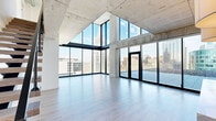 Located at 81 E 14th St in the heart of Kansas City, this two-floor residence showcases a striking industrial-modern architectural design defined by exposed concrete ceilings, structural columns, and expansive floor-to-ceiling glass walls framed in sleek black steel. The open living area on the upper level is anchored by dramatic angled glazing that floods the space with natural light and frames panoramic views of the downtown Kansas City skyline. Light wood-tone flooring runs throughout, complementing the raw concrete overhead and lending warmth to the industrial aesthetic.

A glass-railed mezzanine level overlooks the city below, offering an additional vantage point across the urban landscape. The kitchen is outfitted with high-gloss cabinetry, a substantial waterfall-edge stone island with veined marble-style countertops, stainless steel appliances, a built-in wine cooler, and a marble slab backsplash. Bedrooms feature floor-to-ceiling windows with city and highway views, light flooring, and clean white walls. Bathrooms are finished with large-format marble-look tile, glass-enclosed showers, soaking tub options, and modern floating vanities. The overall ambiance is contemporary, light-filled, and distinctly urban.