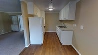 This single-floor space features a galley-style kitchen that serves as its centerpiece. The kitchen is designed for efficient use of space, with white upper and lower cabinetry providing ample storage along both walls. A granite-look countertop runs along one side, housing a stainless steel sink with a modern faucet. The white refrigerator and built-in appliances, including what appears to be a stacked washer and dryer unit, are neatly integrated into the layout. Warm-toned hardwood-style flooring runs throughout, adding a sense of continuity and warmth to the space. Crown molding along the ceiling adds a subtle architectural detail that elevates the overall finish. Overhead lighting provides consistent illumination throughout the kitchen corridor. The neutral beige walls complement the white cabinetry and wood flooring, creating a cohesive, clean aesthetic. The layout opens toward additional living areas, suggesting a practical flow between spaces. Overall, the space presents a tidy, move-in-ready environment suited for everyday living.