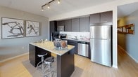 This thoughtfully designed single-floor residence blends modern aesthetics with functional living. The open-concept living area serves as the heart of the space, anchored by large windows that frame urban skyline views and flood the room with natural light. A striking geometric chandelier adds an artistic focal point overhead, while light hardwood flooring ties the space together seamlessly.

The kitchen is outfitted with dark cabinetry, stainless steel appliances, a subway tile backsplash, and a granite-topped island with bar seating — ideal for both casual dining and meal preparation. Track lighting above illuminates the workspace efficiently.

The bedroom is a refined retreat, distinguished by a dramatic marbled accent wall adorned with sculptural globe wall art finished in gold and white. Large windows continue the theme of natural light and outward views, while plush carpeting adds warmth underfoot.

The full bathroom features a sleek, elongated vanity with dark cabinetry and a white countertop, complemented by large-format stone-look tile flooring and a full tub-shower combination. Neutral tones throughout create a calm, spa-like atmosphere.

Overall, the residence presents a cohesive modern design language with curated details, quality finishes, and a bright, airy ambiance enhanced by urban views.
