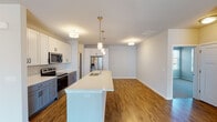 This well-appointed single-floor residence offers a thoughtfully designed open-concept layout that seamlessly connects the kitchen and living areas. The kitchen is a standout feature, boasting a large central island with a white quartz countertop and an integrated sink, two-tone cabinetry with white upper cabinets and slate-blue lower cabinets, and a full suite of stainless steel appliances including a refrigerator with ice and water dispenser, electric range, and built-in microwave. Pendant lighting with gold accents hangs above the island, complementing the warm-toned hardwood-style flooring that extends throughout the main living areas. The open living space offers generous room for furnishings and benefits from a neutral color palette that enhances the bright, airy atmosphere. The bedrooms feature plush carpeting and multiple windows that invite abundant natural light, with views of the surrounding neighborhood. The unit includes two bathrooms: one with a glass-enclosed walk-in shower and wood-look flooring, and a second full bathroom with a shower-tub combination and a slate-blue vanity with a white basin. A dedicated in-unit laundry area with a washer and additional storage closets adds to the practical appeal of this modern, move-in-ready space.