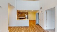 This two-story residence showcases a striking architectural design highlighted by dramatic double-height ceilings in the main living area, which flood the space with natural light through tall windows and sliding glass doors. The ground floor centers around an open-concept layout where the living area flows seamlessly into a well-appointed kitchen featuring warm wood cabinetry, granite countertops, a breakfast bar peninsula with pendant lighting, and a full suite of stainless steel appliances. A second, separate kitchen is also present, outfitted with contemporary light wood cabinetry, matching stainless steel appliances, and clean subway tile backsplash — offering exceptional flexibility. Light hardwood flooring runs throughout the main level, contributing to the bright, airy atmosphere. The upper floor features a loft-style landing that overlooks the living area below, reinforcing the home's sense of openness and vertical scale. Bedrooms are carpeted for comfort and bathed in natural light through windows with blinds, while a spacious walk-in closet provides generous storage. The bathroom is finished with granite countertops, tile flooring, and a full tub-shower combination. Throughout both floors, the clean white walls and recessed lighting create a cohesive, modern ambiance.
