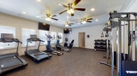 This fitness center offers a comprehensive workout environment designed to support a wide range of exercise routines. The space is finished with durable rubber flooring throughout, providing a safe and practical surface for all types of training. Crown molding and clean white walls lend a polished look to the interior, while multiple windows fitted with horizontal blinds allow natural light to fill the room and offer views to the outside.

Overhead, multiple ceiling fans with integrated lighting ensure consistent air circulation and comfort during workouts. Recessed lighting supplements the natural light, keeping the space bright and well-illuminated. Wall-mounted flat-screen televisions are positioned throughout the room, allowing members to stay entertained during cardio sessions.

The cardio section is well-appointed with treadmills, elliptical trainers, and stationary and recumbent bikes, arranged to maximize floor space and ease of use. A dedicated free weights area features a three-tier dumbbell rack stocked with a full range of weights, accompanied by a adjustable workout bench for strength training. Functional training accessories include medicine balls on a storage rack, stability balls, and suspension training straps mounted to the wall. A yoga mat and additional accessories round out the space, supporting stretching and flexibility work. A secondary door provides convenient exterior access.