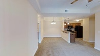 This single-story apartment at Academy Park Apartments offers an open-concept layout that seamlessly connects the living, dining, and kitchen areas. The living and dining spaces are finished with plush wall-to-wall carpeting in a neutral beige tone, creating a warm and inviting atmosphere. A decorative chandelier with candelabra-style lights serves as a focal point in the dining area, adding a touch of elegance to the space. The tray ceiling detail above the dining area adds architectural interest and a sense of height to the room. A ceiling fan is also present, providing both comfort and functionality. The kitchen is partially separated by a breakfast bar with granite countertops and pendant lighting overhead, offering a casual seating option. Light wood cabinetry lines the kitchen walls, complemented by black appliances including a gas range and refrigerator. A HVAC vent is integrated into the ceiling, ensuring year-round climate control. The neutral color palette throughout creates a cohesive and move-in ready environment. A hallway visible from the main living area leads to additional rooms within the unit.