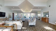 This single-story community clubhouse at 3601 W Broadway in Columbia, Missouri, offers a thoughtfully designed collection of amenity spaces that blend comfort and functionality. The centerpiece is a generously proportioned lounge featuring a striking vaulted, coffered ceiling that draws the eye upward and creates a sense of grandeur. Soft blue-gray walls, patterned carpeting, and an array of stylish seating arrangements — including plush sofas and accent chairs — contribute to a welcoming, refined atmosphere. Floor-to-ceiling windows and glass doors line the perimeter, flooding the space with abundant natural light and providing sweeping views of the surrounding landscaped grounds. A separate dining area offers an intimate setting with a warm wood table, upholstered chairs, crown molding, and curated artwork. The kitchenette is equipped with cabinetry, countertop workspace, appliances, and bar-height seating, ideal for gatherings. The well-appointed fitness center features a full suite of strength and cardio equipment bathed in natural light from large windows overlooking lush greenery. A dedicated business nook provides a computer workstation for residents' convenience. Throughout, the design maintains a cohesive coastal-inspired palette, creating a serene and polished environment.
