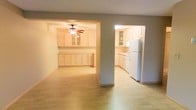 This single-floor space offers a functional and open layout, with interconnected living and kitchen areas that create a cohesive flow. Light-toned hardwood-style flooring runs throughout, lending warmth and continuity across the space.

The main living area is accessed through an open archway and is equipped with a ceiling fan, providing both light and air circulation. Extensive built-in cabinetry lines the walls, featuring light wood finishes and glass-front upper cabinets, offering generous storage while maintaining an airy, uncluttered aesthetic. The neutral, taupe-toned walls complement the cabinetry and flooring.

Adjacent to the main area, the kitchen is accessible through a separate doorway and continues the same light wood cabinetry theme. It features ample counter space, additional upper and lower cabinetry with glass-front accents, and includes a full-size white refrigerator. The kitchen layout is well-organized and practical, providing sufficient workspace for everyday use.

Overall, the space maintains a clean, understated aesthetic with consistent finishes throughout, making it a versatile and functional environment.
