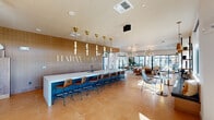 Located at 2500 East Marco Polo Road in Phoenix, AZ, this single-floor clubhouse for Harwood on Cave Creek offers a generously sized, open-concept gathering space combining a dining area and living room. The interior showcases a contemporary design aesthetic with warm wood-paneled walls, large-format tile flooring, and light hardwood accents throughout.

A striking centerpiece of the space is the long communal bar topped with a white stone countertop and accented by a bold blue base, lined with leather barstools and illuminated by a series of gold pendant lights suspended from the ceiling. The branded Harwood signage on the wood-paneled back wall adds a polished, professional identity to the area.

The living room section features a built-in linear fireplace beneath a wall-mounted television, flanked by open wood shelving with curated décor. Seating arrangements include leather sofas, woven accent chairs, and patterned rugs that add texture and warmth. A sculptural gold branching chandelier serves as a dramatic focal point overhead.

Floor-to-ceiling black-framed glass doors and expansive windows flood the interior with natural light while offering views of the surrounding desert landscaping and palm trees. The dining area benefits from large sliding glass doors, sheer drapery, and lush potted tropical plants that bring a refreshing organic element indoors. The overall ambiance is modern, inviting, and sophisticatedly curated.