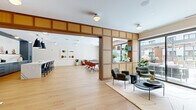 Located at 1105 Pleasant St in Oak Park, Illinois, this thoughtfully designed single-floor amenity space offers a seamless blend of functionality and modern aesthetics. Light oak hardwood flooring flows throughout, anchoring a warm, contemporary palette of natural wood tones, soft neutrals, and muted blues.

The open-plan layout accommodates several distinct zones. A comfortable lounge area features plush, rounded green armchairs arranged around a low coffee table on a sculptural area rug, bathed in natural light from large sliding glass doors that open directly onto a furnished outdoor terrace. The terrace, visible through floor-to-ceiling glazing, is equipped with pergola-covered seating, extending the living space outdoors.

A striking kitchen and bar area commands attention with its elongated marble-topped island lined with cane-backed counter stools, complemented by slate-blue cabinetry, a marble slab backsplash, open wood shelving, and bold black pendant lighting overhead.

Adjacent to the kitchen, a flex dining and gathering area provides long communal tables and colorful accent chairs, ideal for group events or casual meetings. Warm wood-paneled dividers with upper display shelving elegantly partition zones while maintaining visual openness. A separate flex room with rich brown floor-to-ceiling drapery offers an adaptable space for private events. Curated artwork is displayed throughout, lending the space a gallery-like refinement.