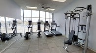 This fitness center is designed to support a well-rounded workout experience within a single-floor layout. The space is defined by its striking floor-to-ceiling windows that span an entire wall, flooding the interior with abundant natural light and offering open views of the surrounding exterior. A ceiling fan overhead provides comfortable air circulation throughout the room.

The flooring consists of durable rubber tile, a practical and impact-resistant surface well-suited for exercise equipment and high-traffic use. Cardio equipment is prominently featured, including multiple treadmills and an elliptical trainer, each unit equipped with individual display screens. A stationary cycle and an ab bench add further variety to the cardio and core training options available.

On the strength training side, a multi-station cable machine offers a range of resistance exercises, and a set of free weights rounds out the equipment selection. The light gray walls and drop ceiling with recessed lighting contribute to a clean, functional aesthetic that keeps the focus on fitness. The overall ambiance is bright, open, and purpose-built, making this space a convenient and fully equipped facility for daily exercise routines.