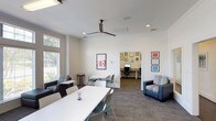 The Study Lounge is a thoughtfully designed, single-floor space built to accommodate a range of academic needs. The main area features multiple white collaborative tables paired with teal and white chairs, providing ample room for group study sessions. Ceiling fans overhead ensure comfortable airflow throughout. Large windows with transom panels line the perimeter, flooding the space with natural light and offering views of the surrounding outdoor landscape. Adjustable blinds allow for light control when needed.

Comfortable seating nooks are integrated throughout, each featuring upholstered armchairs with plaid accents, ottomans, and side tables — ideal for independent reading or casual study. Movable privacy panels offer flexible semi-private workstations within the open floor plan, catering to those who prefer a quieter environment without full enclosure.

A dedicated computer lab, accessible directly from the main lounge, features a granite-topped desk spanning the room's width, housing multiple desktop computers and a printer. The walls are decorated with an eclectic gallery arrangement of framed artwork and motivational prints, adding personality to the functional space. Patterned carpet throughout ties the modern, clean aesthetic together, making this lounge a productive and welcoming environment available around the clock.