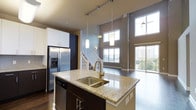 This two-story residence is defined by its dramatic double-height ceilings in the main living area, creating a sense of openness and grandeur. The open-concept layout seamlessly connects the kitchen and living space, making it ideal for both daily living and entertaining.

The kitchen is well-appointed with granite countertops, dark cabinetry, and a large central island featuring a double undermount sink with a brass-finish pull-down faucet. Pendant lighting on a track rail hangs above the island, providing focused illumination. Stainless steel appliances, including a French door refrigerator with an in-door ice and water dispenser, complement the modern aesthetic. White upper cabinetry contrasts the darker lower cabinets, adding visual balance.

The adjacent living area benefits from floor-to-ceiling windows and a sliding glass door that opens to a balcony, flooding the space with natural light and offering views of the surrounding cityscape. Dark hardwood flooring runs throughout, adding warmth and continuity. A staircase with white balusters leads to the second floor, reinforcing the home's two-level design. The overall ambiance is modern, airy, and refined.
