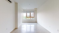 This single-floor space showcases a clean, contemporary design defined by polished concrete floors that run throughout and crisp white walls that enhance the sense of openness and light. The open-plan layout connects the kitchen and living area seamlessly, creating a fluid and functional flow. Large wood-framed windows at the far end of the living area flood the space with natural light while offering views of the surrounding urban landscape, including greenery and neighboring buildings.

The kitchen is well-appointed with light wood cabinetry extending from floor to ceiling, providing generous storage. Stainless steel appliances include a built-in wall oven, a bottom-freezer refrigerator, a dishwasher, and a gas cooktop. Dark countertops and a deep blue-toned backsplash add contrast to the warm cabinetry tones. Open shelving offers additional storage and display space, while a stainless steel sink completes the functional workspace.

The bathroom features classic white subway tile surrounding a full soaking tub and shower, complemented by a curved chrome curtain rod, a wood-accented vanity with a dark countertop, and the same polished concrete flooring found throughout. A wall-mounted mini-split unit provides climate control. The overall ambiance is minimalist, bright, and urban-modern.