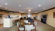 This single-floor multipurpose common area is thoughtfully designed to accommodate a variety of activities and social interactions. The space is organized into several distinct functional zones that flow seamlessly into one another. At the forefront is a round dining table surrounded by upholstered chairs, creating an inviting spot for casual meals or group gatherings. Adjacent to this is a comfortable lounge area furnished with dark upholstered sofas and armchairs, ideal for relaxed conversation. Along one wall, a bar-height counter with padded stools provides an additional casual seating option. A dedicated workstation area with a desk and office chair is tucked along the perimeter, offering a quiet space for productivity. The interior is finished with warm beige walls accented by a contrasting dark crown molding detail that adds a touch of architectural character. Recessed lighting and dual ceiling fans ensure the space remains well-lit and comfortable throughout. Carpeted flooring in a neutral tone ties the entire room together, while natural light filters in through windows at the far end, brightening the overall ambiance. The layout and furnishings make this a versatile and functional communal environment.