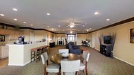 This single-floor multipurpose common area is thoughtfully designed to accommodate a variety of activities and social interactions. The space is organized into several distinct functional zones that flow seamlessly into one another. At the forefront is a round dining table surrounded by upholstered chairs, creating an inviting spot for casual meals or group gatherings. Adjacent to this is a comfortable lounge area furnished with dark upholstered sofas and armchairs, ideal for relaxed conversation. Along one wall, a bar-height counter with padded stools provides an additional casual seating option. A dedicated workstation area with a desk and office chair is tucked along the perimeter, offering a quiet space for productivity. The interior is finished with warm beige walls accented by a contrasting dark crown molding detail that adds a touch of architectural character. Recessed lighting and dual ceiling fans ensure the space remains well-lit and comfortable throughout. Carpeted flooring in a neutral tone ties the entire room together, while natural light filters in through windows at the far end, brightening the overall ambiance. The layout and furnishings make this a versatile and functional communal environment.