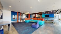 Located in Memphis, TN, this dynamic single-floor clubhouse delivers a bold and energetic atmosphere through its striking use of color, modern industrial design, and thoughtfully defined activity zones. Polished concrete floors extend throughout the entire space, reflecting light and providing a sleek industrial foundation that contrasts beautifully with the vivid orange, turquoise, and cobalt blue accents found throughout.

The main lounge area anchors the space with a large modular sectional sofa in turquoise and orange, complemented by a statement purple accent chair and area rugs. Adjacent to the lounge, a game zone features a foosball table set against a dramatic sunburst-patterned blue mural wall with mounted flat-screen TVs and high-top bar seating.

Wooden slatted ceiling details and exposed ductwork add an industrial-chic character, while recessed and pendant lighting ensure the space is well-lit throughout. A row of upholstered banquette booths with stone tile backdrops offers a cozy setting for casual dining or conversation.

Glass-walled conference rooms provide transparent, professional workspaces with direct views to the outdoors through expansive floor-to-ceiling windows. Open co-working areas with modern tables and chairs round out the collaborative offerings. The space is a well-curated blend of social, recreational, and productive environments.
