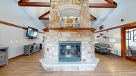 Located at Falcon Glen in Greenfield, Wisconsin, this single-floor community clubhouse offers a generously sized, open-concept gathering space designed with both comfort and character in mind. The centerpiece of the main lounge is a striking floor-to-ceiling natural stone fireplace, anchored by a warm wood mantel and rising dramatically toward soaring vaulted ceilings accented with exposed dark wood beam trusses. The expansive great room flows seamlessly across light-toned hardwood-style flooring, providing ample room for multiple seating arrangements, including a leather lounge area and media wall with a mounted flat-screen television. Large windows along the perimeter allow natural light to fill the space, complementing the bright white walls and rich wood trim throughout. A set of elegant glass-paneled French doors separates the main lounge from an adjacent dining area, which features multiple tables and chairs suited for casual gatherings or community events. Windows and glass-paned exterior doors in the dining area provide direct views of and access to an outdoor pool area. Decorative aviation-themed wall art adds a distinctive, playful touch to the overall aesthetic, reinforcing the Falcon Glen identity throughout the space.