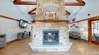 Located at Falcon Glen in Greenfield, Wisconsin, this single-floor community clubhouse offers a generously sized, open-concept gathering space designed with both comfort and character in mind. The centerpiece of the main lounge is a striking floor-to-ceiling natural stone fireplace, anchored by a warm wood mantel and rising dramatically toward soaring vaulted ceilings accented with exposed dark wood beam trusses. The expansive great room flows seamlessly across light-toned hardwood-style flooring, providing ample room for multiple seating arrangements, including a leather lounge area and media wall with a mounted flat-screen television. Large windows along the perimeter allow natural light to fill the space, complementing the bright white walls and rich wood trim throughout. A set of elegant glass-paneled French doors separates the main lounge from an adjacent dining area, which features multiple tables and chairs suited for casual gatherings or community events. Windows and glass-paned exterior doors in the dining area provide direct views of and access to an outdoor pool area. Decorative aviation-themed wall art adds a distinctive, playful touch to the overall aesthetic, reinforcing the Falcon Glen identity throughout the space.