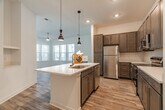 This single-story, three-bedroom, two-bathroom home in Collierville, Tennessee offers a well-designed open floor plan with a cohesive blend of modern finishes and functional living spaces. The combined kitchen and living area serves as the heart of the home, where a central island with a undermount sink and pendant lighting anchors the kitchen. Rich dark-toned cabinetry, a subway tile backsplash, stainless steel appliances, and light wood-style flooring complete the kitchen's polished look. The adjacent living room receives generous natural light through a bank of large windows with blinds and features a ceiling fan and a glass-paned door providing outdoor access. The three bedrooms are carpeted for comfort, with the secondary bedrooms featuring bright windows with views of the surrounding area. Each bedroom includes closet space, and the primary suite is served by a spacious en-suite bathroom with a double-sink vanity, dark cabinetry with brushed nickel hardware, wood-style flooring, and a glass-enclosed tiled shower. A dedicated laundry room houses a full-size washer and dryer. Three closets provide ample storage throughout, and a connecting hallway ties the layout together efficiently.