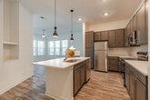 This single-story, three-bedroom, two-bathroom home in Collierville, Tennessee offers a well-designed open floor plan with a cohesive blend of modern finishes and functional living spaces. The combined kitchen and living area serves as the heart of the home, where a central island with a undermount sink and pendant lighting anchors the kitchen. Rich dark-toned cabinetry, a subway tile backsplash, stainless steel appliances, and light wood-style flooring complete the kitchen's polished look. The adjacent living room receives generous natural light through a bank of large windows with blinds and features a ceiling fan and a glass-paned door providing outdoor access. The three bedrooms are carpeted for comfort, with the secondary bedrooms featuring bright windows with views of the surrounding area. Each bedroom includes closet space, and the primary suite is served by a spacious en-suite bathroom with a double-sink vanity, dark cabinetry with brushed nickel hardware, wood-style flooring, and a glass-enclosed tiled shower. A dedicated laundry room houses a full-size washer and dryer. Three closets provide ample storage throughout, and a connecting hallway ties the layout together efficiently.