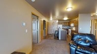 This single-floor space features an open layout that connects the living and kitchen areas into one cohesive, functional zone. The living area is finished with wall-to-wall carpet in a neutral tone, providing a comfortable foundation, while the kitchen and surrounding hallway transition to dark tile flooring, clearly delineating the two spaces.

The kitchen is well-appointed with medium oak-toned cabinetry spanning both upper and lower sections, offering ample storage. A central island with a built-in countertop and bar stool seating serves as both a prep space and casual dining area. White appliances, including a refrigerator, electric range, and built-in microwave, are neatly integrated into the layout. A pendant light fixture hangs above the island, complementing the flush-mount ceiling lights found throughout.

A hallway visible from the main living area leads to multiple rooms, each accessible through classic six-panel doors with light wood trim that coordinates with the cabinetry. The overall color palette — warm beige walls, dark ceilings, and honey-toned wood accents — creates a cozy, grounded atmosphere throughout the space.