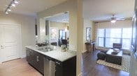 This single-floor residence showcases a thoughtfully designed open-concept layout that promotes a sense of spaciousness and connectivity between living areas. The kitchen serves as a central hub, featuring striking dark cabinetry paired with light granite countertops, a stainless steel sink, and a dishwasher. Pendant lighting hangs elegantly above the kitchen area, adding both functionality and visual warmth to the space.

A structural column subtly defines the transition between the kitchen and the adjoining living areas without compromising the open flow. The living area is finished with rich dark hardwood flooring, creating a warm contrast against the neutral beige walls. Natural light pours in generously through multiple windows fitted with blinds and curtains, brightening each zone of the home.

Beyond the kitchen pass-through, a bedroom is visible, bathed in natural light from its own windows and furnished with complementary décor. The overall ambiance of the space is modern and inviting, with a cohesive neutral color palette, layered lighting, and quality finishes that contribute to a polished, contemporary aesthetic throughout.
