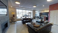 This single-floor space offers a well-appointed and inviting communal environment designed for both relaxation and social interaction. The lounge area is anchored by a pair of rich, dark leather Chesterfield sofas and a contemporary chaise lounge, arranged around a glass-top coffee table atop a plush area rug. Decorative accents, including a guitar wall mount and a colorful accent stool, lend the space a casual, eclectic personality.

Large, floor-to-ceiling windows along the front wall invite generous amounts of natural light, creating a bright and open atmosphere while offering clear views of the surrounding streetscape. The drop-panel ceiling is fitted with recessed flush-mount lighting and track lighting, ensuring the space is well-lit throughout.

Adjacent to the seating area is a kitchenette and bar space, complete with dark cabinetry, a raised countertop with barstool seating, and practical amenities that make it suitable for gatherings and events. The overall design blends modern furnishings with warm, welcoming touches, resulting in a functional yet comfortable communal space ideal for residents or guests to gather, unwind, and connect.