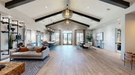 Located at 622 Brigham Rd in Greensboro, NC, this single-floor community space offers a thoughtfully designed interior that blends modern aesthetics with warm, inviting details. The centerpiece of the space is a dramatic vaulted ceiling adorned with bold dark exposed beams, complemented by striking wire-frame pendant chandeliers that serve as sculptural focal points throughout.

Wide-plank, wood-look flooring runs continuously across the entire floor, unifying the various functional zones. The open layout encompasses multiple seating arrangements, including a comfortable lounge area with a plush sofa, a geometric area rug, and a tile-accented fireplace wall fitted with built-in pipe-style shelving. A steel-framed glass partition provides subtle division between zones while preserving the open, airy feel.

A café-style nook features built-in banquette seating alongside small bistro tables, accented by curated artwork and warm sconce lighting. Toward the rear, a full kitchen with stainless steel appliances, dark cabinetry, and a long bar-height counter serves as an entertainment hub. Large windows and glass French doors flood the space with abundant natural light and provide direct views of and access to an outdoor patio area, seamlessly extending the interior to the outside.