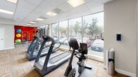 Located at 511 Four Mile Rd in Alexandria, Virginia, this single-floor fitness center is thoughtfully designed to support a well-rounded workout experience. The space features a clean, modern aesthetic with a drop-panel ceiling outfitted with recessed lighting, patterned carpet tile flooring with bold orange accent lines, and a combination of white and vibrant red accent walls that energize the environment.

Large floor-to-ceiling windows line the perimeter, flooding the space with natural light and providing views of the surrounding greenery and streetscape. The cardio zone is well-equipped with multiple treadmills, an elliptical trainer, and a stationary bike, all positioned along the windowed wall to maximize natural light during workouts. A wall-mounted flat-screen television is conveniently positioned for viewing during exercise sessions.

The strength training area includes a multi-function cable machine with an adjustable bench, suitable for a wide range of resistance exercises. A full rack of dumbbells in varying weights provides ample options for free weight training. Full-length mirrors along one wall enhance the sense of space and allow users to monitor their form. Colorful medicine balls are also available, adding a functional training element to the space. The overall design is bright, functional, and well-organized.
